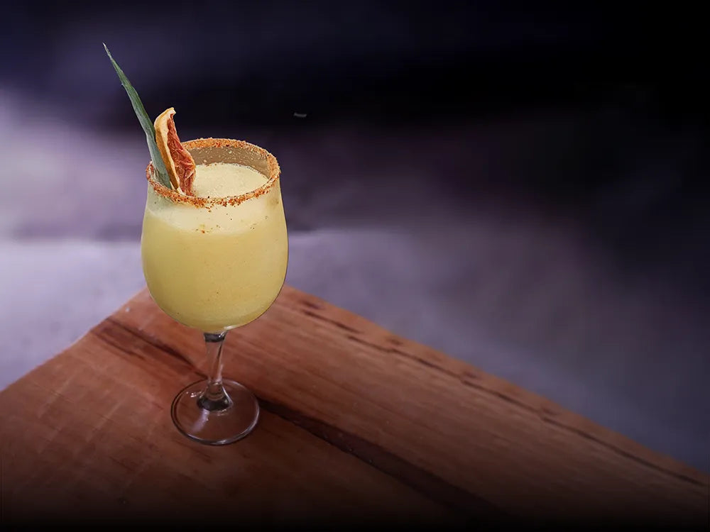 Cóctel Amanecer en Casa Xamu con mezcal artesanal Casa Xamu, borde escarchado, toronja deshidratada y hoja de agave, servido en copa sobre madera.