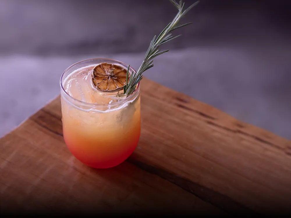Cóctel Puesta de Sol en Matatlán con mezcal artesanal Casa Xamu, rodaja de naranja deshidratada y romero fresco, con degradado de colores naranja y rojo servido en vaso sobre madera