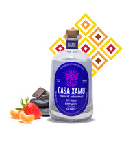 Botella de mezcal Espadín Joven Salvaje de Casa Xamu acompañada de fresas y mandarinas frescas, piedras de río que simbolizan su sabor terroso, barra de cacao, chile piquín y hoja de laurel. Fondo con textura de rombos en colores que representan los sabores sensoriales: frutal, especiado, terroso y picante.