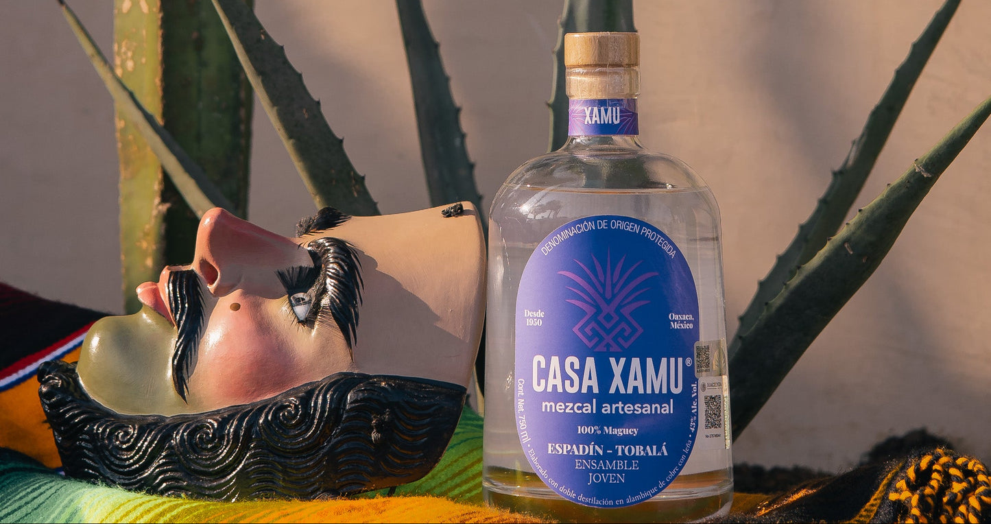 Botella de mezcal artesanal Casa Xamu Ensamble Joven Espadín Tobalá junto a máscara tradicional de madera y agaves, sobre sarape colorido